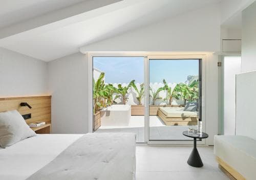 A serene hotel room with a minimalist design opens to a lush balcony adorned with tropical plants, perfect for travelers seeking relaxation and modern comfort.