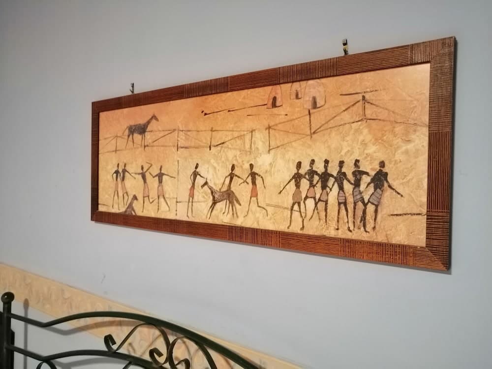 A rustic wall art piece depicting ancient tribal life with figures, animals, and structures in earthy tones, framed elegantly, adds cultural ambiance to an indoor space, ideal for travelers seeking authentic decor inspiration.