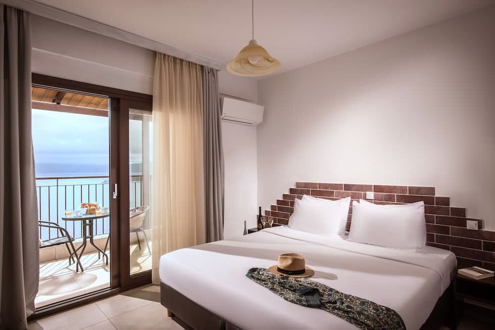 A cozy hotel room with a neatly made bed adorned with a hat and scarf, features a brick accent wall, modern furnishings, and a serene balcony overlooking the sea, ideal for travelers seeking relaxation and comfort.
