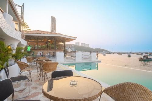 A serene coastal restaurant scene features an infinity pool overlooking the sea, with wicker chairs and tables set for relaxation under a clear sky, ideal for travelers seeking a tranquil seaside dining experience.