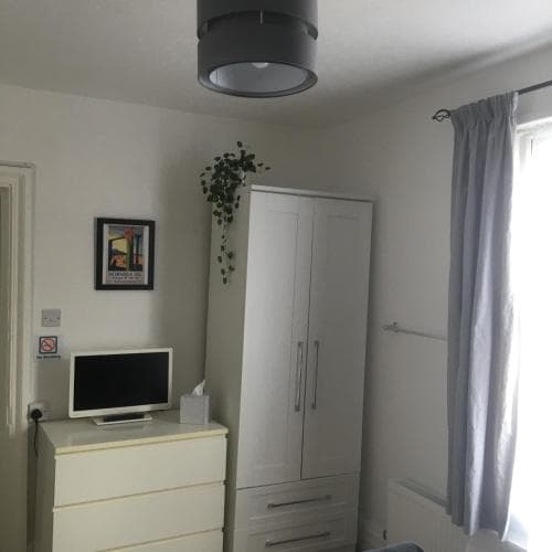 A cozy bedroom with white furniture, a small TV on a dresser, a hanging plant, and soft natural light through a window with gray curtains, ideal for travelers seeking a comfortable and minimalist accommodation.