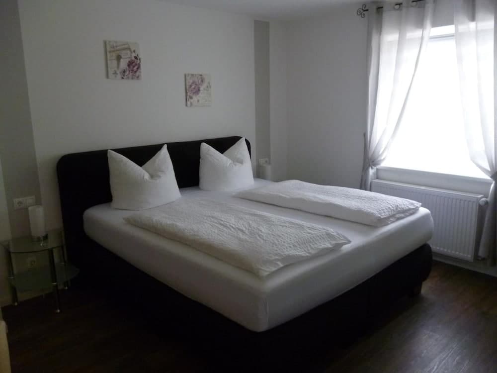 A cozy hotel room features a neatly made double bed with white linens, soft natural light from a window with sheer curtains, and minimalist decor, ideal for travelers seeking comfort and simplicity.