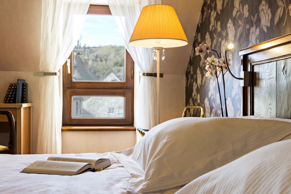A cozy bedroom with floral wallpaper, a warmly lit lamp, and an open book on the bed invites relaxation, perfect for travelers seeking a serene retreat with a view of quaint rooftops outside.