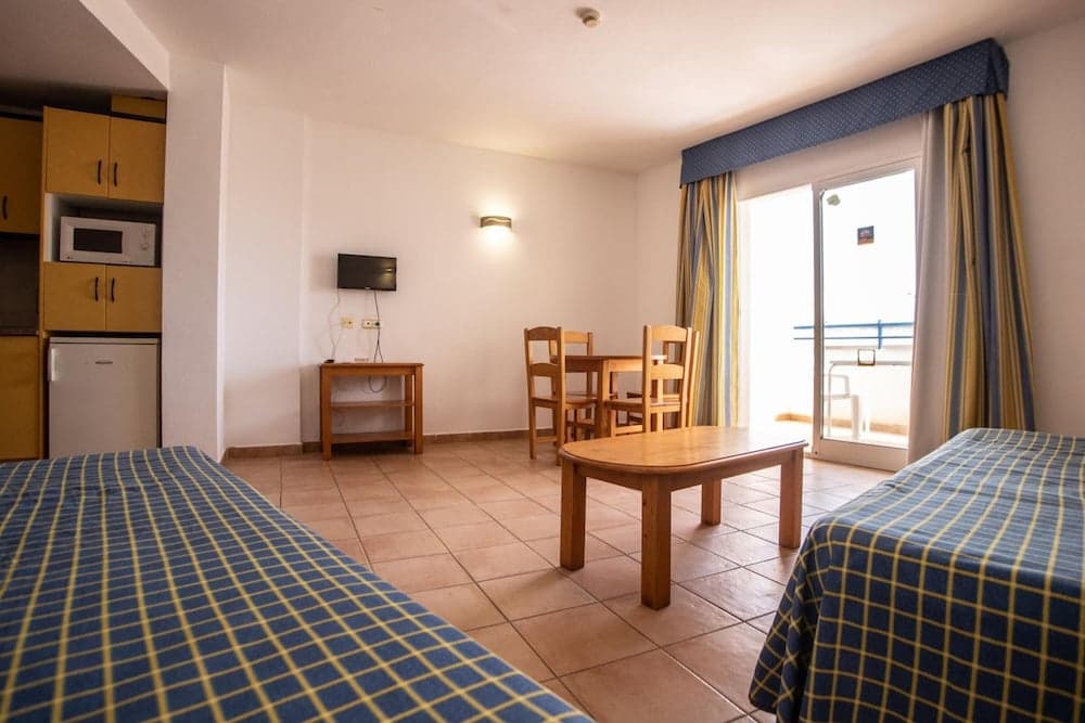 A cozy hotel room with tiled floors, wooden furniture, a kitchenette, and a balcony offers a comfortable stay for travelers seeking relaxation and convenience.