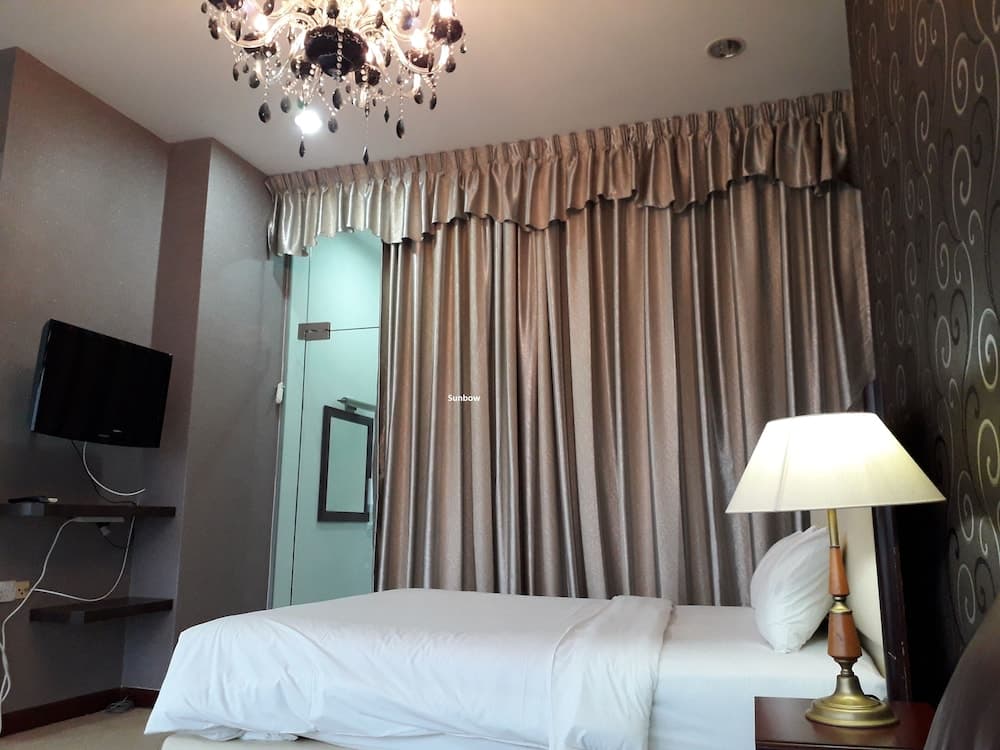 A cozy hotel room features a neatly made bed, elegant curtains, a chandelier, and a mounted TV, creating a comfortable and inviting atmosphere ideal for travelers seeking relaxation.