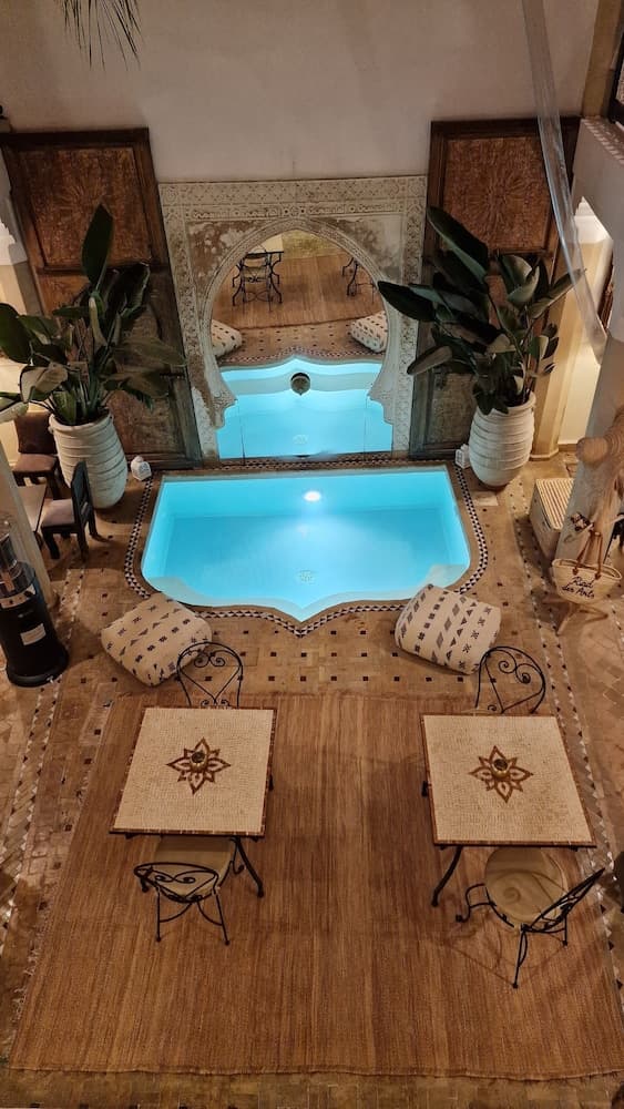 An inviting indoor courtyard features a serene pool surrounded by patterned cushions, ornate tables, lush plants, and traditional decor, creating a tranquil oasis ideal for relaxation and cultural immersion during travel.