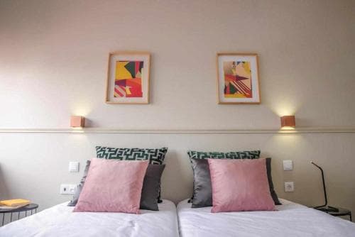 A cozy hotel room features twin beds adorned with pink pillows, geometric-patterned headboards, and abstract art above, creating a modern and inviting atmosphere ideal for travelers seeking comfort and style.