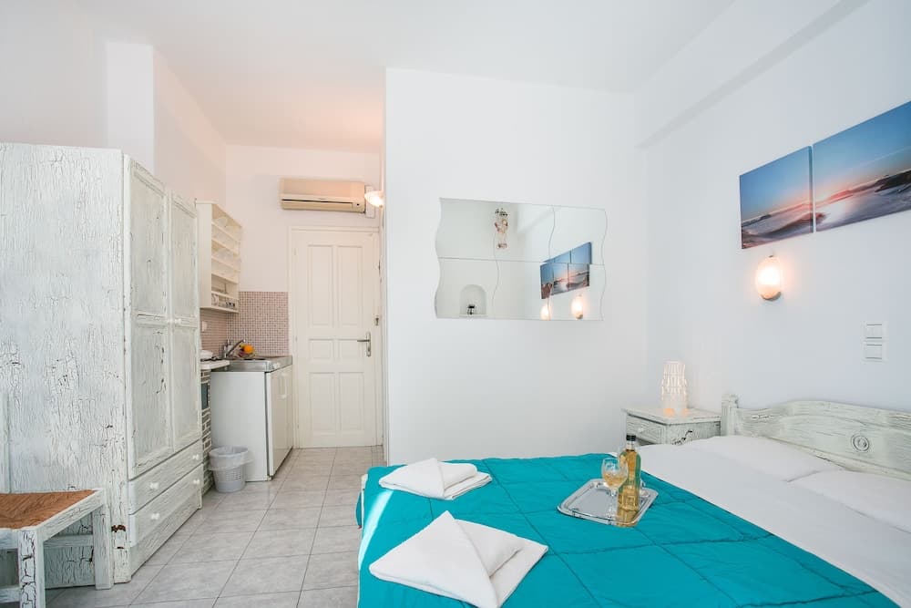 A cozy studio apartment with a bright turquoise bedspread, white rustic furniture, and a compact kitchenette, ideal for a serene beachside getaway.