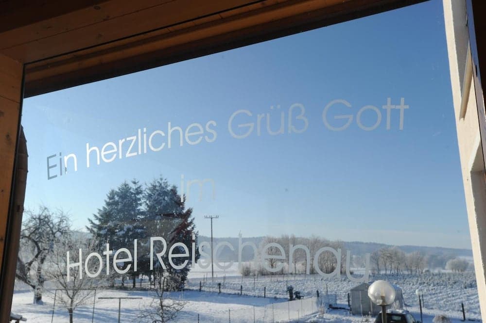 A serene winter landscape viewed through a window with "Ein herzliches Grüß Gott im Hotel Reischenau" written on it, evokes a cozy, inviting atmosphere perfect for a tranquil getaway in a snowy countryside setting.