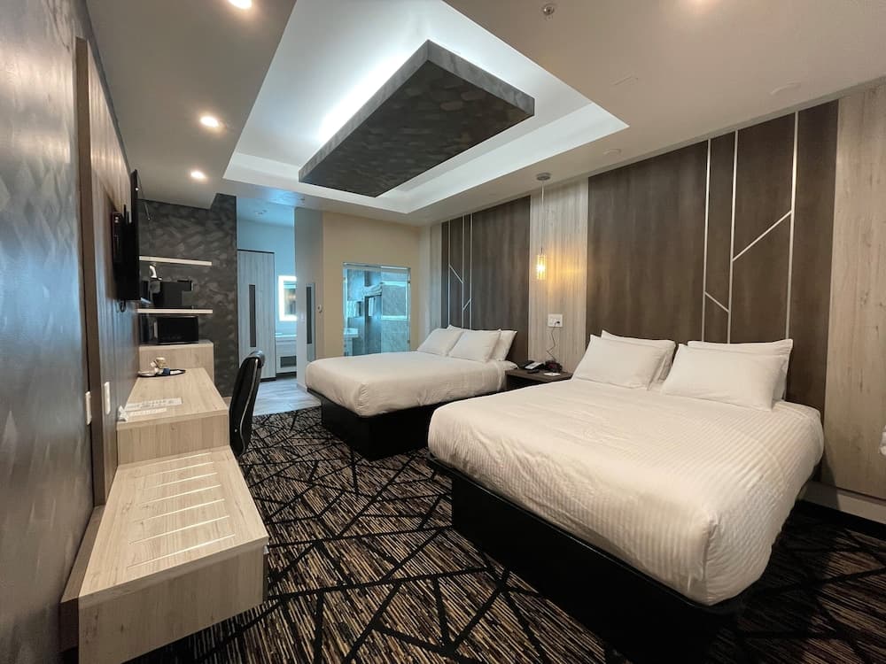 A cozy hotel room features two neatly made beds under a modern geometric ceiling light, complemented by a stylish desk area and a bathroom view, ideal for travelers seeking comfort and contemporary design.