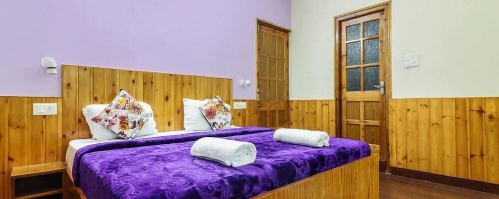 A cozy hotel room features a wooden bed with purple bedding, floral pillows, neatly rolled towels, and warm-toned walls, ideal for travelers seeking comfort and relaxation.