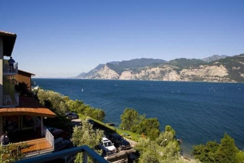 A serene lakeside view with vibrant buildings overlooking the expansive blue waters of Lake Garda, surrounded by lush greenery and distant mountains, ideal for tranquil vacations and scenic photography.