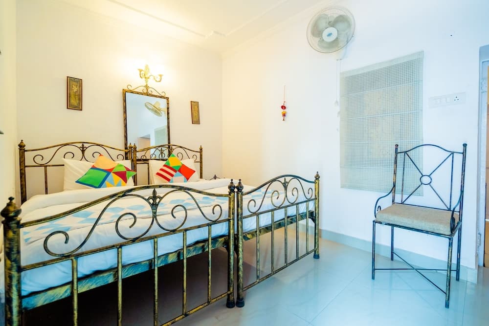 A cozy bedroom with a metal bed adorned with colorful pillows, a cushioned chair, and a wall-mounted fan, ideal for travelers seeking a comfortable and budget-friendly accommodation.