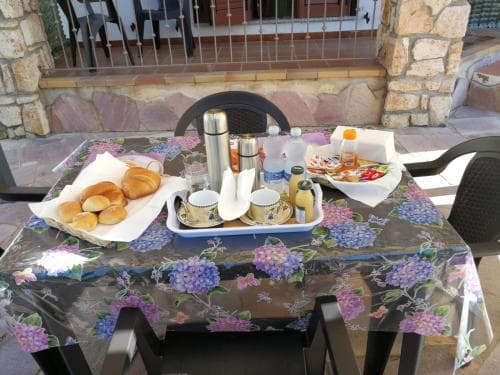 A cozy outdoor breakfast setup on a floral tablecloth, featuring fresh pastries and beverages, invites travelers to enjoy a serene morning in a rustic stone-walled patio setting.