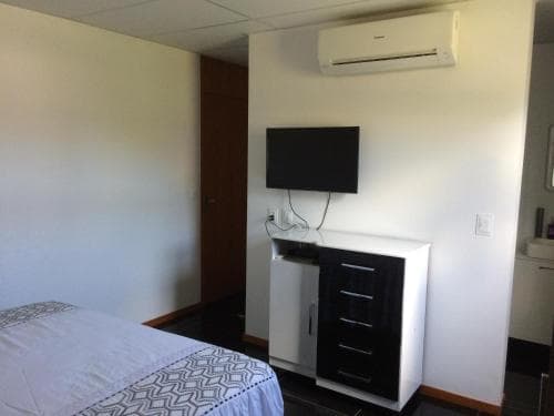 A cozy hotel room features a neatly made bed, a mounted TV above a sleek white cabinet, and an air conditioner, ideal for travelers seeking comfort and convenience.