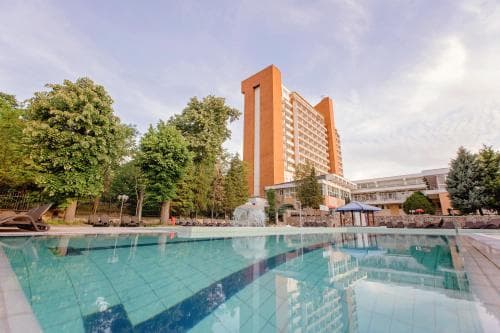 A serene outdoor pool scene with clear blue water reflects the tall orange hotel building amidst lush green trees, ideal for travelers seeking relaxation in a resort setting.