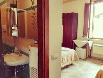 A cozy bedroom with tiled floors, wooden accents, and natural light streaming through a window, paired with an adjacent bathroom featuring a sink and mirror, ideal for travelers seeking a quaint and charming accommodation experience.