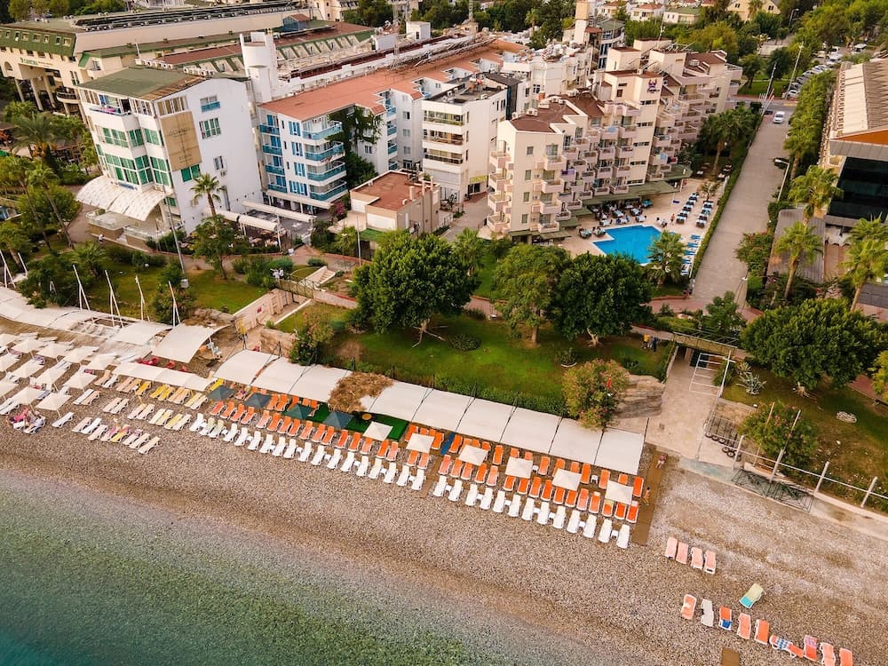 An aerial view captures a vibrant coastal resort scene with colorful beach loungers under white umbrellas on a pebble beach, surrounded by lush greenery and modern apartment buildings, ideal for travelers seeking a relaxing seaside getaway.