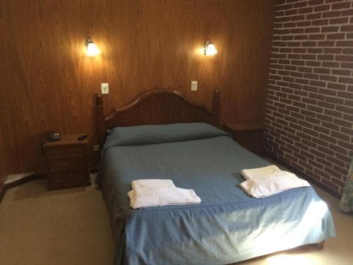 A cozy hotel room features a neatly made bed with blue linens, wooden paneling, brick accent wall, and warm lighting, ideal for travelers seeking comfort and simplicity.