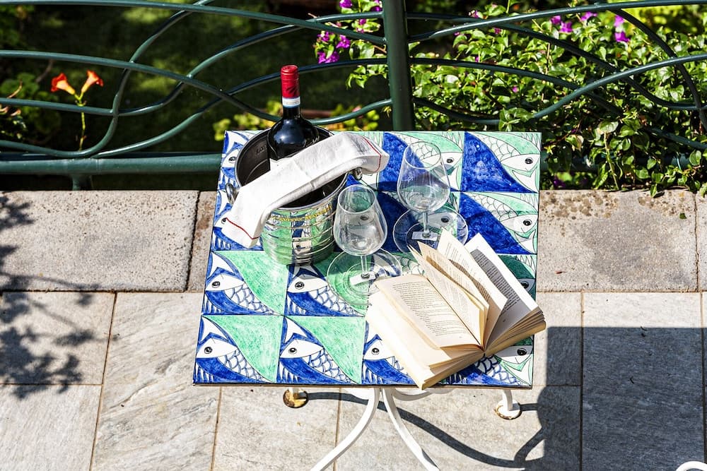 A serene outdoor patio scene features a colorful mosaic table adorned with an open book, wine glasses, a chilled bottle of wine in an ice bucket, and lush greenery in the background, perfect for a relaxing travel getaway.