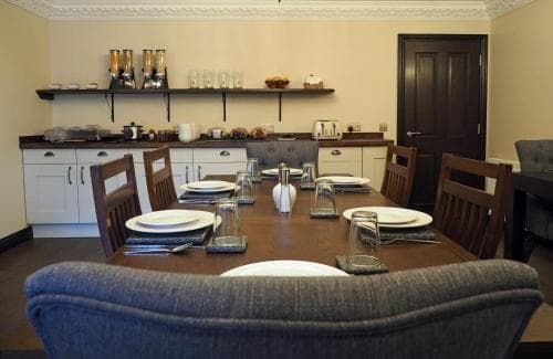 A cozy dining area with a wooden table set for a meal, surrounded by chairs, in a well-lit room with kitchen cabinets and shelves, ideal for travelers seeking a comfortable self-catering accommodation experience.