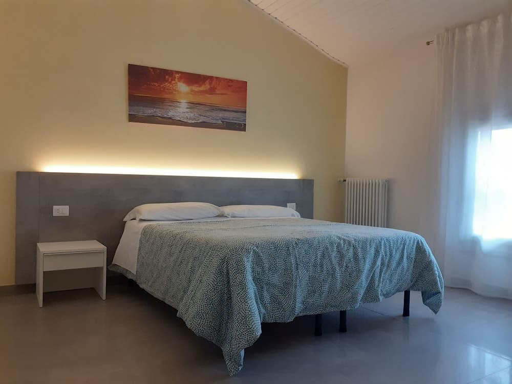 A cozy bedroom with a modern bed setup, featuring a concrete headboard, a serene sunset artwork, soft lighting, and sheer curtains, ideal for travelers seeking a tranquil retreat.