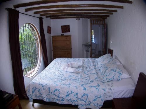 A cozy bedroom with a rustic charm features a neatly made bed under wooden beams, a round window with lush greenery outside, and warm lighting, ideal for travelers seeking a tranquil retreat in a countryside setting.