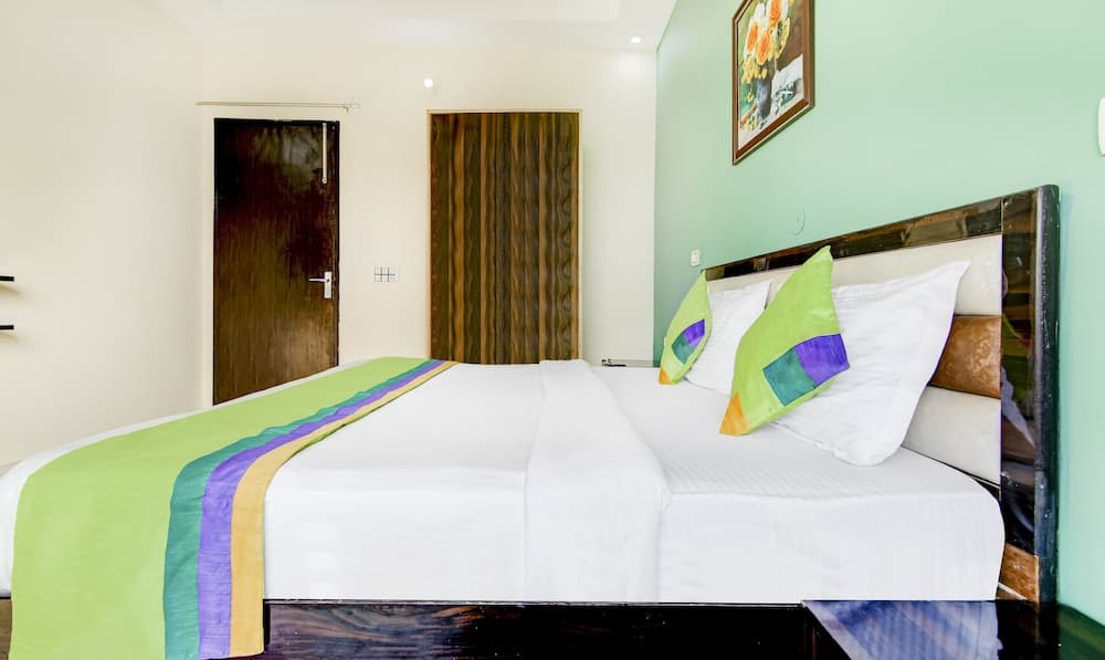 A cozy hotel room with a neatly made bed adorned with colorful pillows and a green throw, set against soft green walls and wooden doors, ideal for travelers seeking comfort and relaxation.