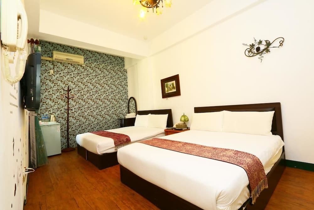 A cozy hotel room features two neatly made beds with white linens and red accents, hardwood floors, a vintage chandelier, and a patterned accent wall, ideal for travelers seeking comfort and charm.
