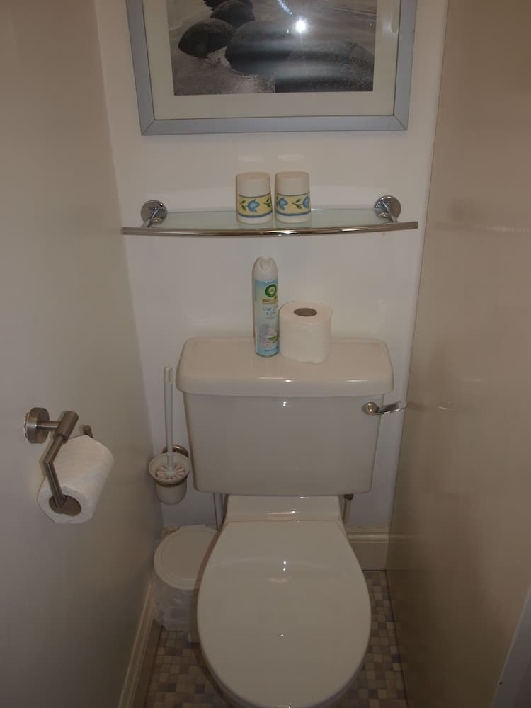 A cozy bathroom corner features a toilet with a glass shelf above holding decorative cups and a framed beach scene, ideal for travelers seeking a clean and organized restroom experience.