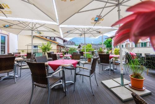 A cozy outdoor terrace with wooden flooring, vibrant red tables, wicker chairs under white umbrellas, and lush greenery offers a serene dining spot with mountain views, perfect for travelers seeking relaxation in an alpine setting.