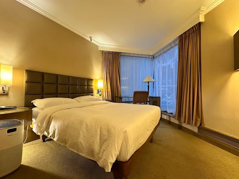 A cozy hotel room at The Kowloon Hotel in Hong Kong features a neatly made bed, warm lighting, and a large window with curtains, ideal for travelers seeking comfort and relaxation.