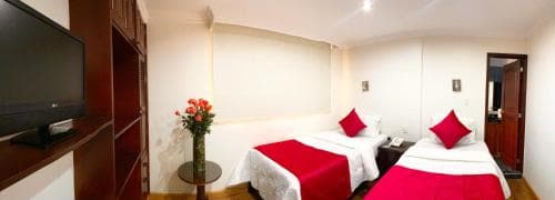 A cozy hotel room features two neatly made beds with red accents, a flat-screen TV on a wooden cabinet, and a vase of flowers on a side table, ideal for travelers seeking comfort and simplicity.