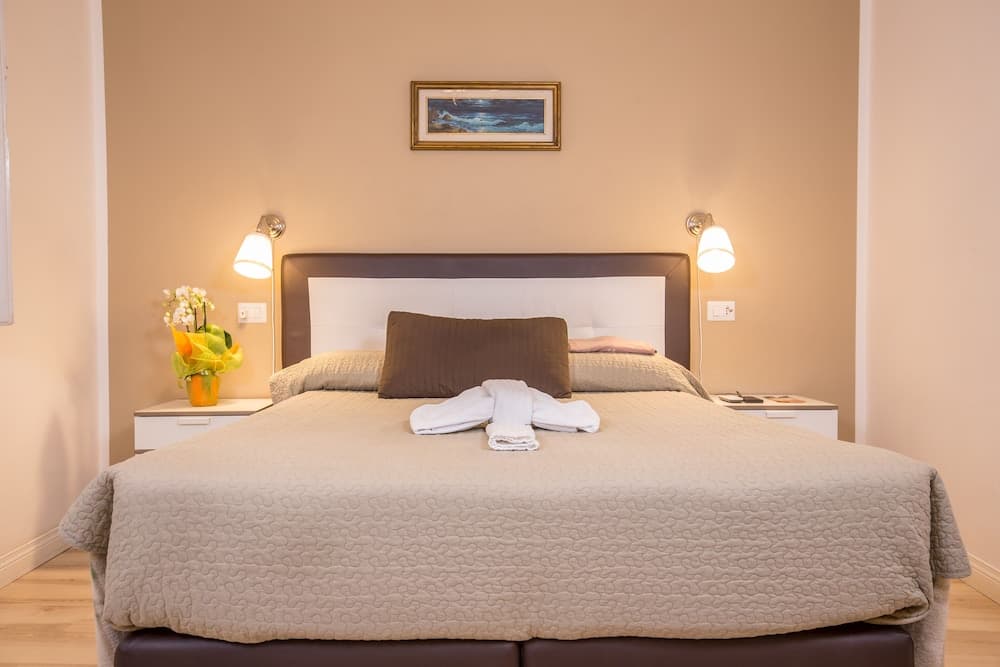 A cozy hotel bedroom features a neatly made bed with soft lighting, wall-mounted lamps, a small vase of flowers on the nightstand, and a serene painting above the headboard, ideal for travelers seeking comfort and relaxation.