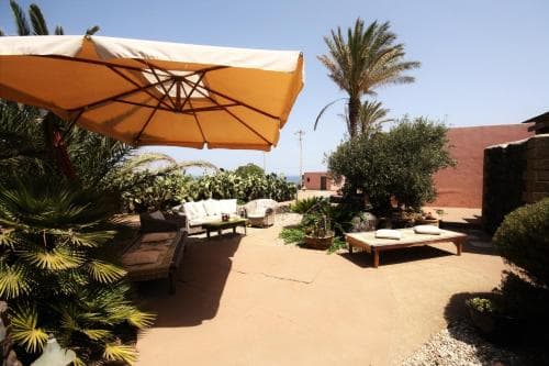 A serene outdoor patio with comfortable seating under a large orange umbrella, surrounded by lush greenery and palm trees, offers a tranquil retreat ideal for relaxation during a beachside getaway.