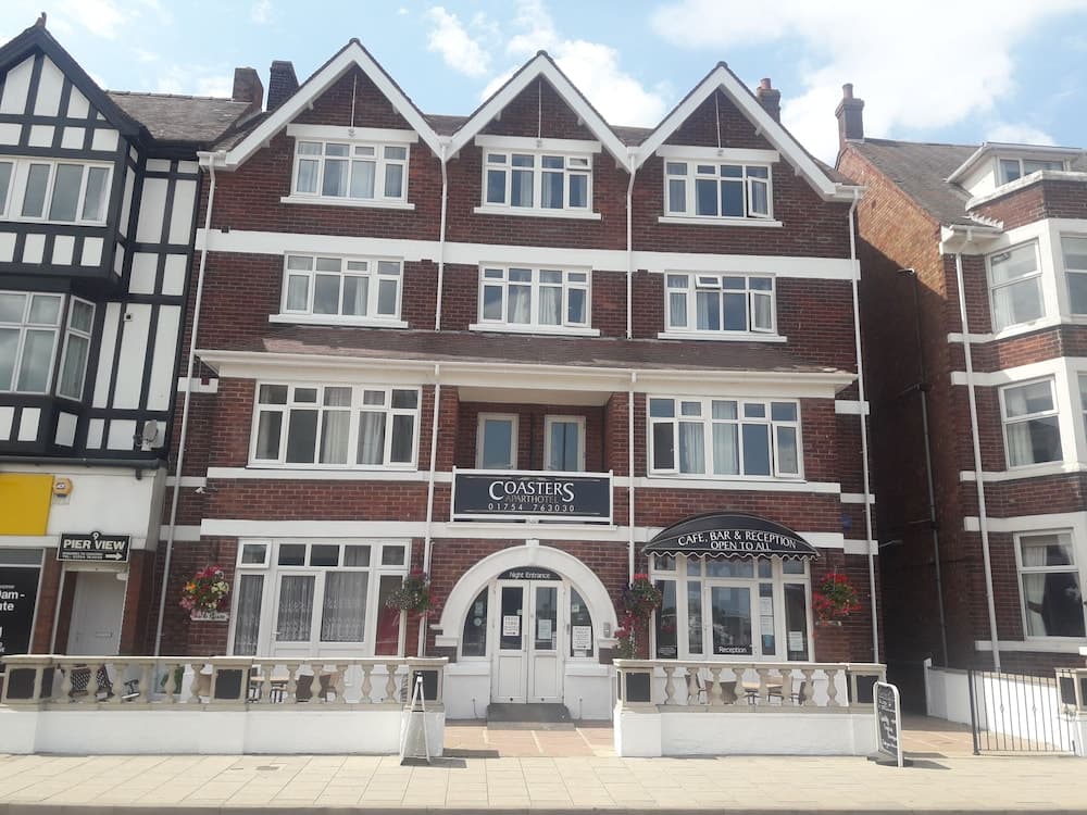 A charming brick building with white trim houses Coasters Cafe, Bar & Reception under a sunny sky, ideal for travelers seeking a cozy seaside retreat with welcoming signage and floral accents.