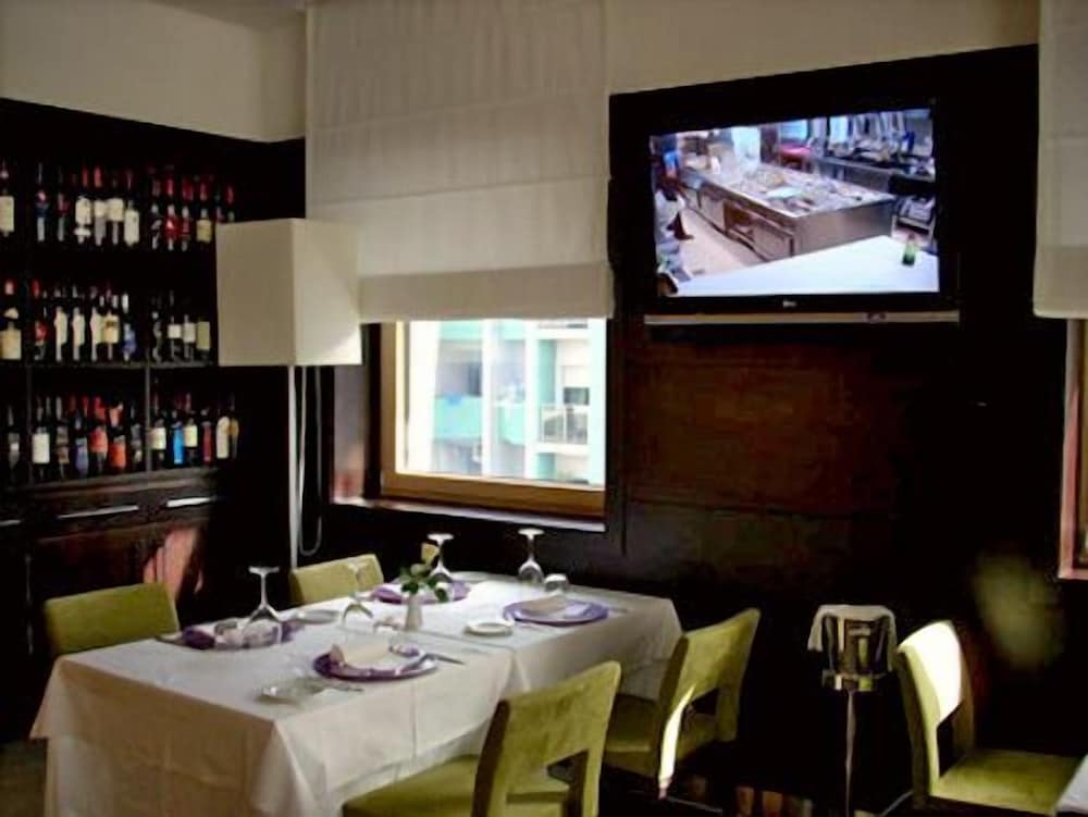 A cozy restaurant dining area features a neatly set table by the window, a wall-mounted TV showing kitchen activity, and a wine rack, ideal for travelers seeking an intimate dining experience.