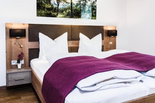 A cozy hotel room features a neatly made bed with a purple throw blanket, wooden bedside tables, and a forest-themed wall art, creating a serene and inviting atmosphere ideal for travelers seeking relaxation.