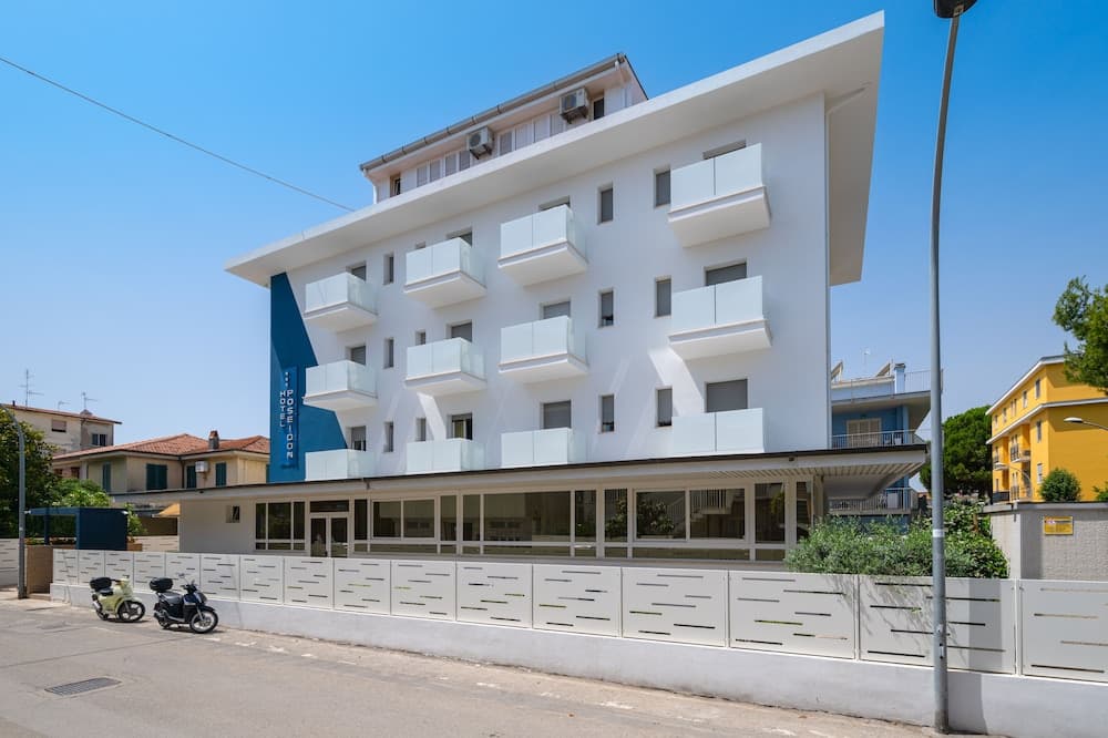 A modern white hotel with blue accents stands under a clear blue sky, featuring balconies and a welcoming entrance, ideal for travelers seeking a bright and stylish accommodation in an urban setting.