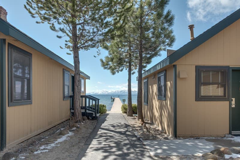 A serene lakeside pathway lined with cozy cabins and towering pine trees leads to a tranquil dock overlooking snow-capped mountains, ideal for a peaceful retreat at the Franciscan Lakeside Lodge in Tahoe Vista, CA.