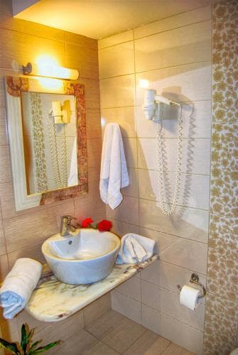 A cozy hotel bathroom features warm lighting, tiled walls, a marble sink countertop adorned with red flowers, and neatly arranged towels, ideal for travelers seeking comfort and cleanliness.