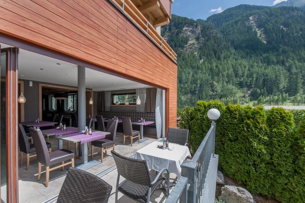 A cozy mountain lodge terrace with purple tables, wicker chairs, and lush greenery offers a serene dining spot for travelers seeking relaxation amidst breathtaking alpine views.