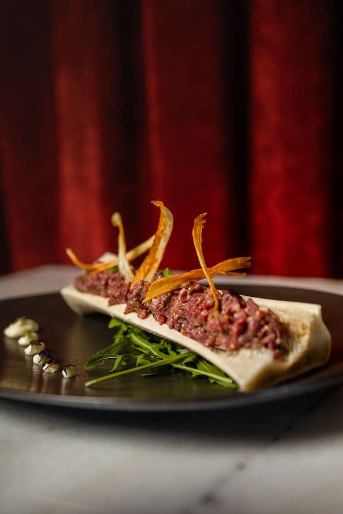 A gourmet beef tartare dish artfully presented on a sleek plate against a rich red curtain backdrop, evoking an upscale dining experience perfect for food enthusiasts exploring fine cuisine destinations.