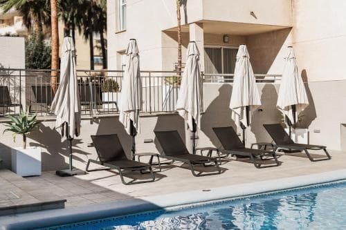 A serene hotel patio features a sparkling pool lined with lounge chairs and closed umbrellas, ideal for relaxing under the sun in a tropical resort setting.