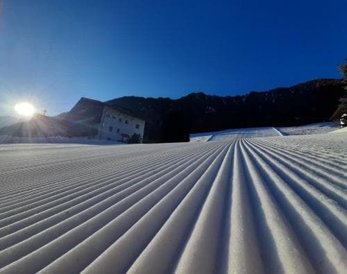 A serene ski resort scene features freshly groomed snow tracks under a clear blue sky, with a quaint building nestled against the backdrop of majestic mountains, ideal for winter sports enthusiasts seeking a peaceful getaway.
