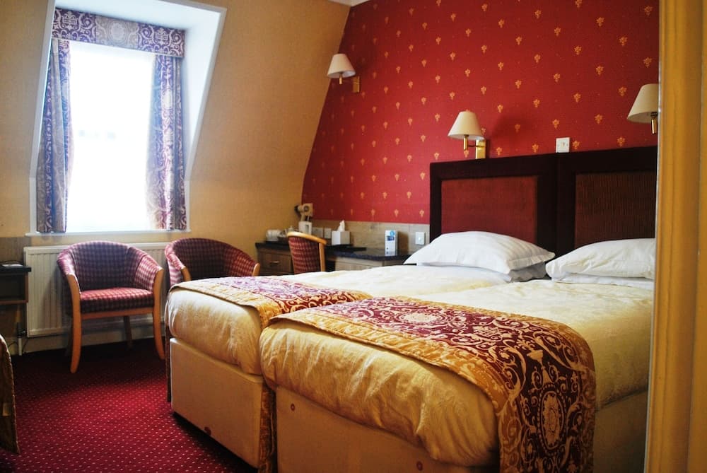 A cozy hotel room features twin beds adorned with red and gold linens, a warm red wall, wooden chairs by a bright window, and bedside lamps, ideal for travelers seeking comfort and charm.