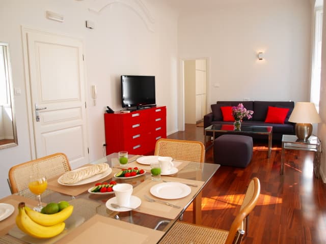 A cozy apartment in Dubrovnik features a bright dining area with fresh fruit, a modern living room with a red cabinet and black sofa, hardwood floors, and natural light, ideal for travelers seeking comfort and style.