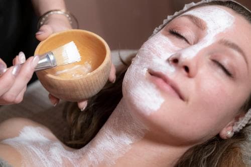 A serene spa scene captures a woman receiving a facial mask treatment, with a wooden bowl and brush in hand, evoking relaxation and self-care for wellness travelers.