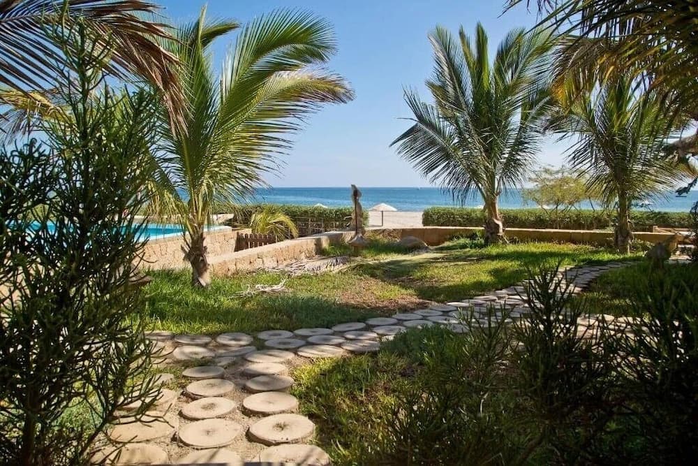A serene tropical resort scene features a stone pathway winding through lush greenery and palm trees, leading to a tranquil beach and ocean view, ideal for vacationers seeking relaxation and natural beauty.