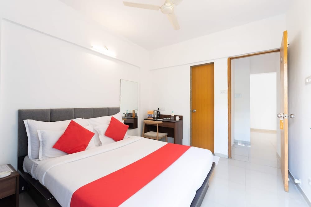 A cozy hotel room features a neatly made bed with red accents, a small desk area, and bright white walls, ideal for travelers seeking comfort and simplicity.